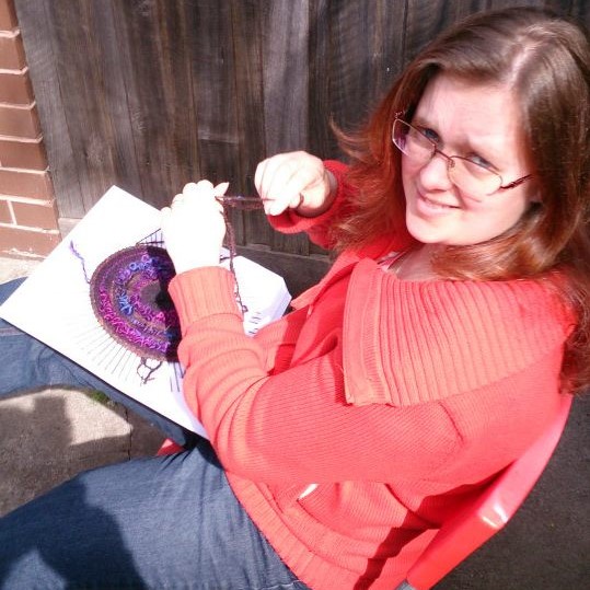 A woman sitting outside in the sun holding yarn and a square of cardboard. The cardboard has a circle of woven yarn in the centre of it.