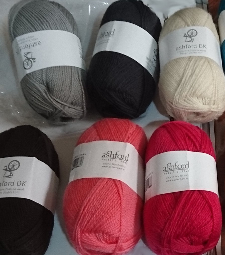 6 balls of Ashford DK wool yarn, from top left: smoke (light grey), granite (dark grey), natural white, chocolate (dark brown), coral (light pink) and rogue (dark pink. 