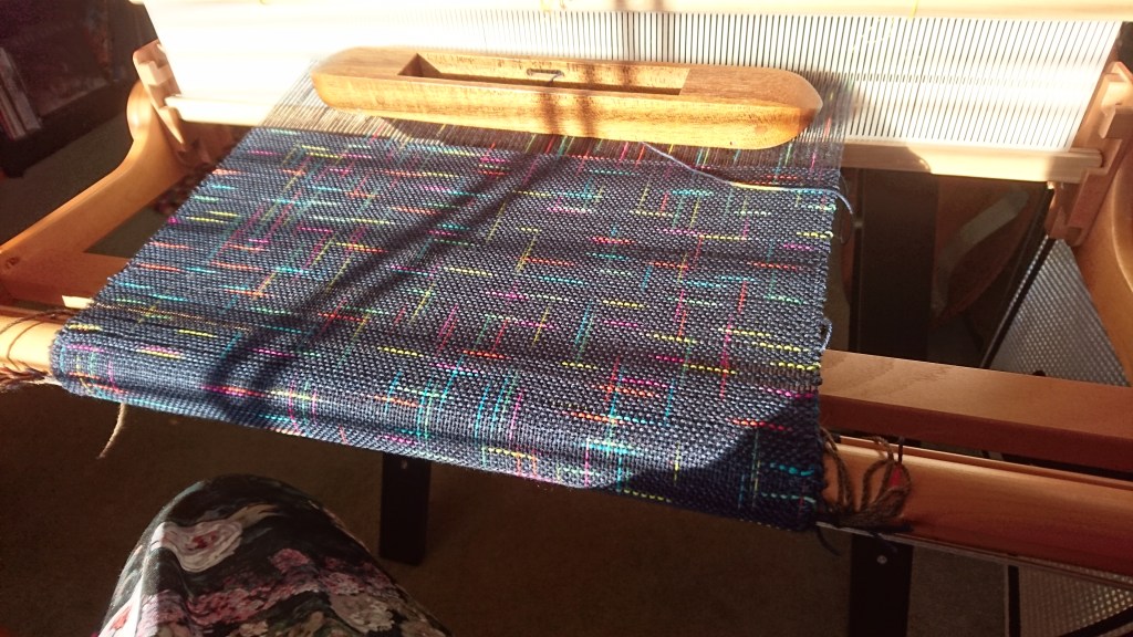 Rigid heddle loom with weaving in progress, with bright sunlight shining on the surface of the weaving. The yarn in both warp and weft is dark blue with random short stripes of green, yellow, pink, light blue and orange. The stripes intersect each other at right angles like an abstract geometric or grid pattern.