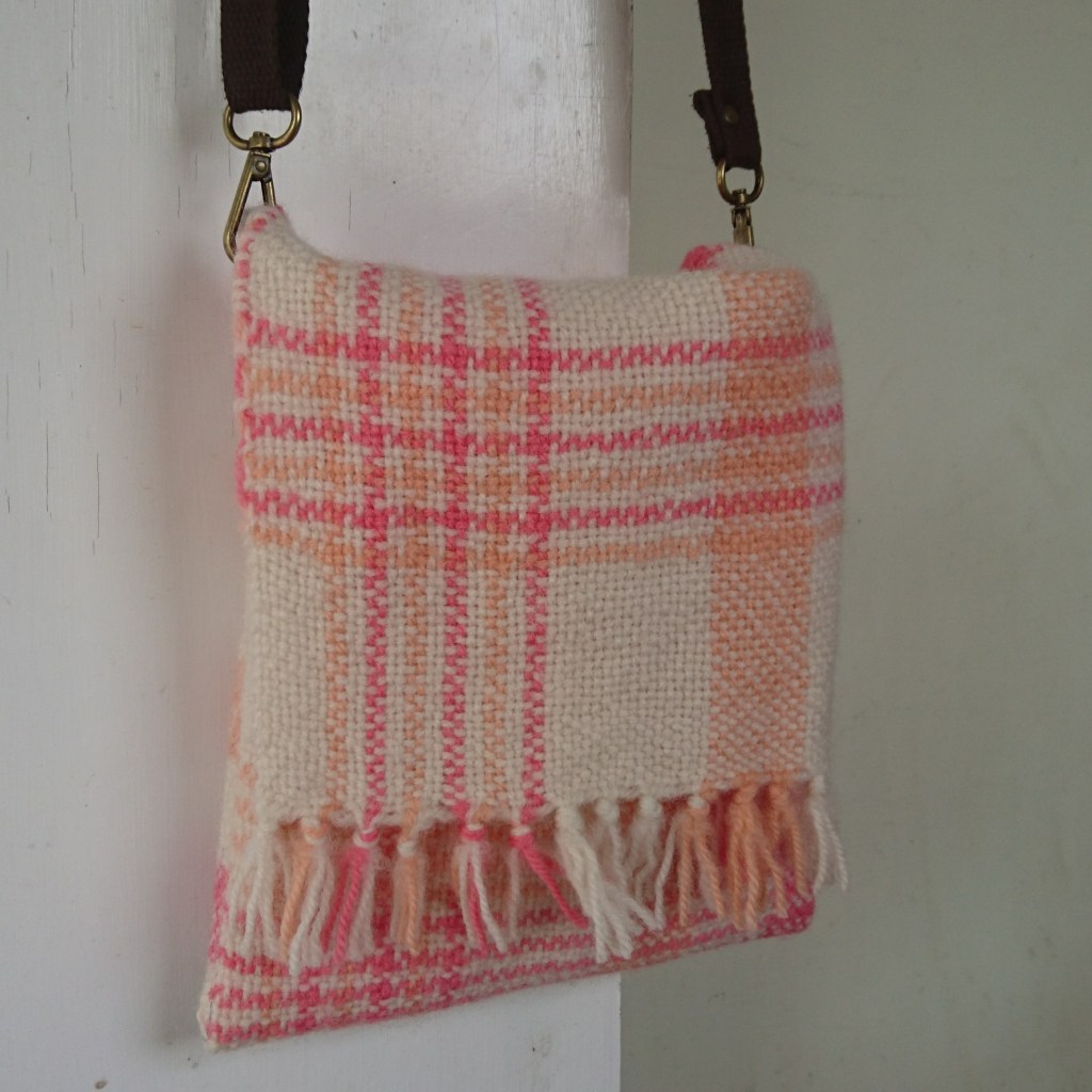 Small square messenger / satchel bag made of woven cloth in white with peach and pink stripes and checks, with a short fringe in the front flap. It's hanging from a brown strap with metal clips on a white door