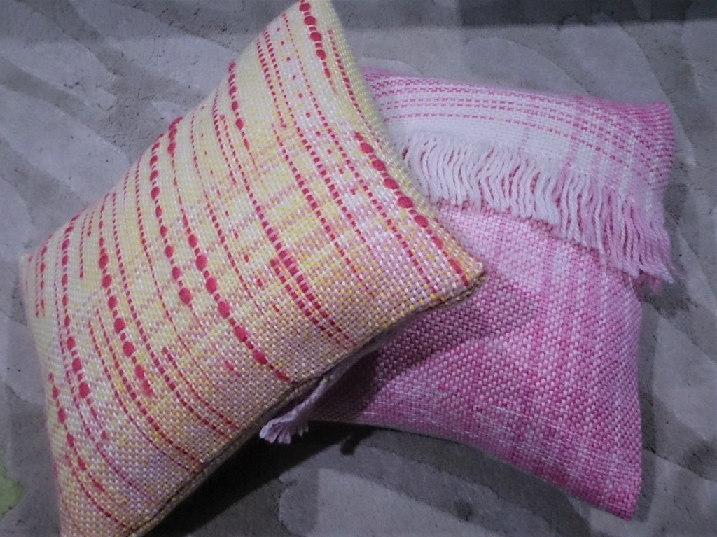 Two handwoven cushions on a white & grey rug. The left cushion is yellow and pink, with bright pink streaks and dots of texture, the right cushion is pink and white with a fringe about 2/3 of the way up.