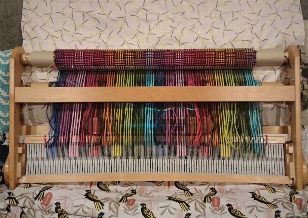 A folded knitters loom resting on patterned fabric (wattle blooms and black cockatoos). There is a project in-progress on the loom composed of bright rainbow stripes and the woven cloth as it winds onto the front beam shows a log-cabin pattern.