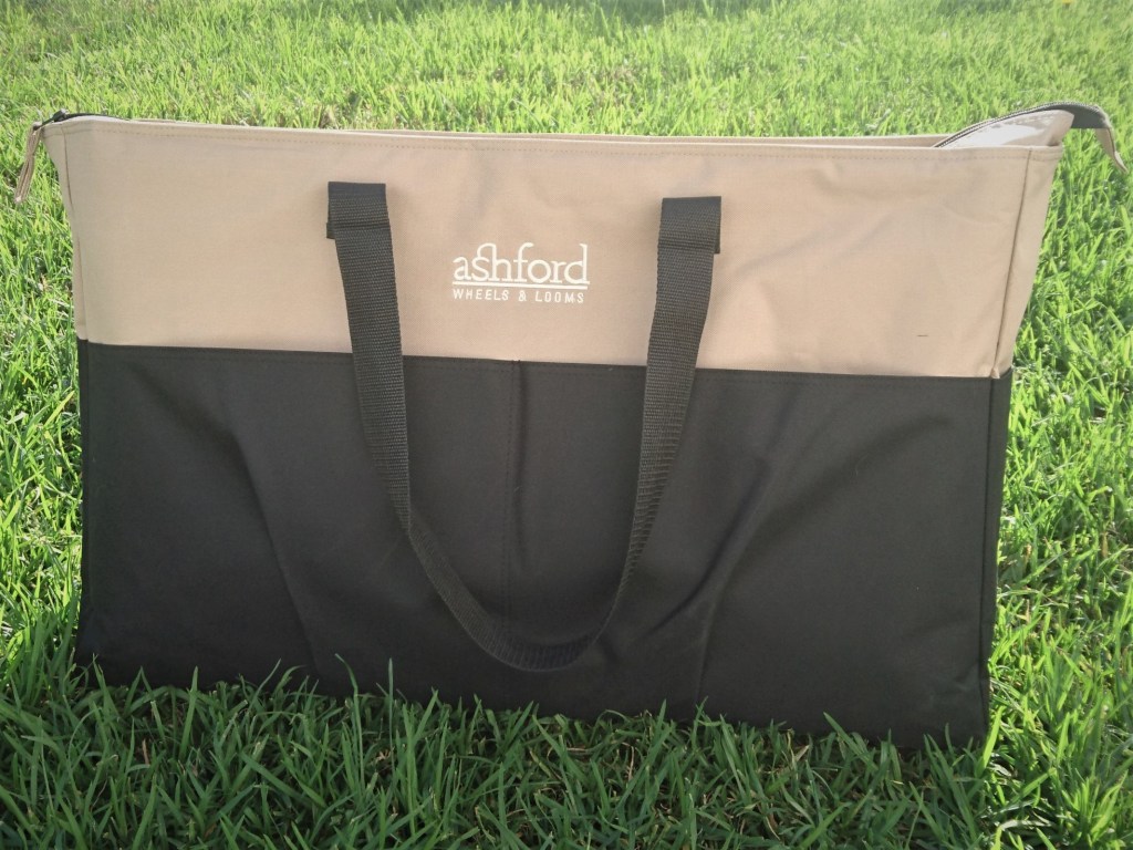 Large beige and black bag sitting on the grass, with a black carry strap and the words Ashford Wheels & Looms embroidered on in white.