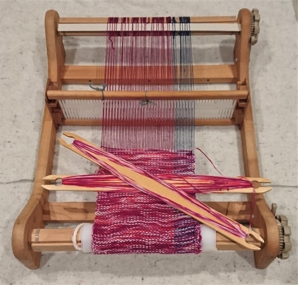 Rigid Heddle loom sitting on white fabric with a raspberry and blue warp and variegated pink & white weft. Two stick shuttles rest on the weaving.