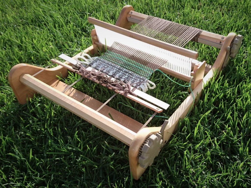 Rigid Heddle loom sitting on green grass. There is a brown warp on the loom with a little bit of white and green yarn just started weaving.