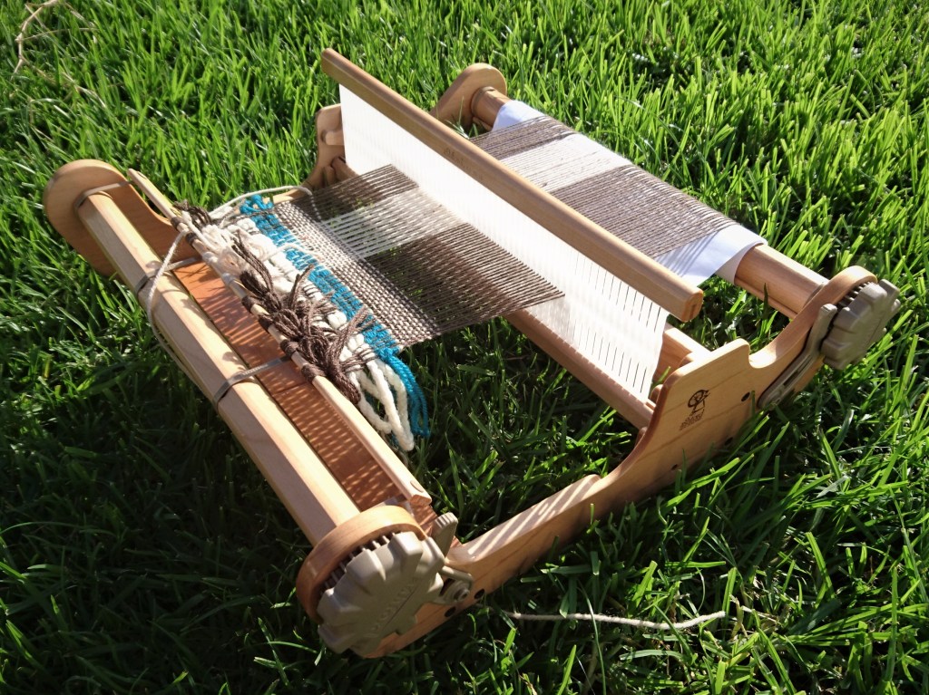 SampleIt loom sitting on green grass. There is a brown & white warp on the loom with a little bit of white and blue yarn just started weaving.