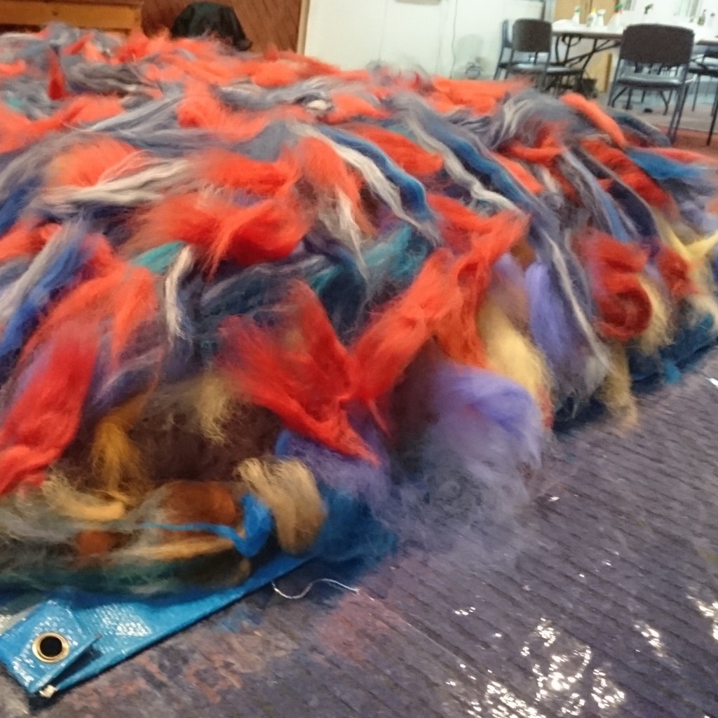 a large pile of fluffy tufts of fibre (wool, alpaca, silk, flax etc) in many colours, spread out over a blue tarp and plastic covers, there are lots of chairs & tables in the background.