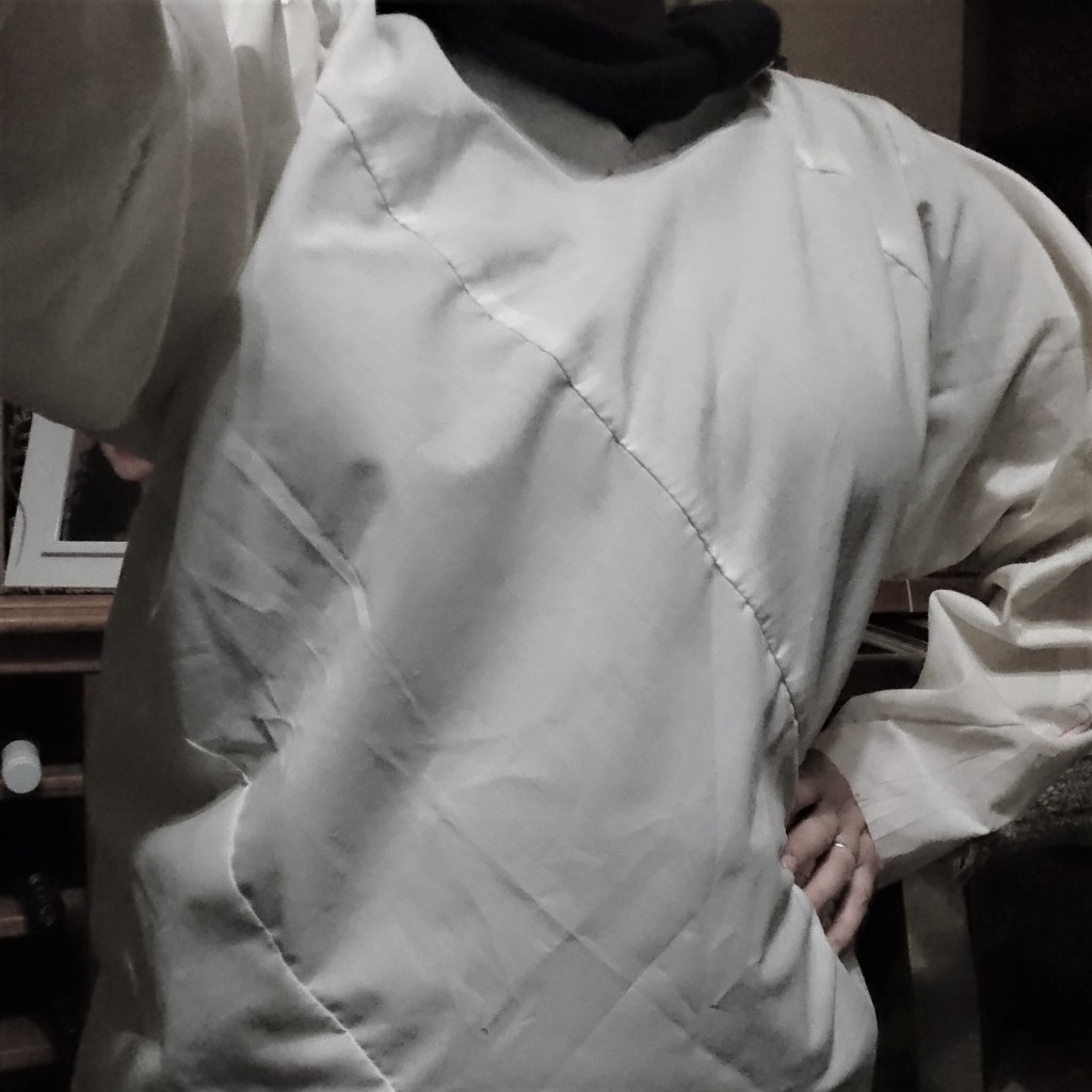 A cropped photo showing only my torso and arms, wearing a plain white long-sleeved cotton top with diagonal seams running across the body & arms, one hand outstretched holding the camera and hand on my hip.