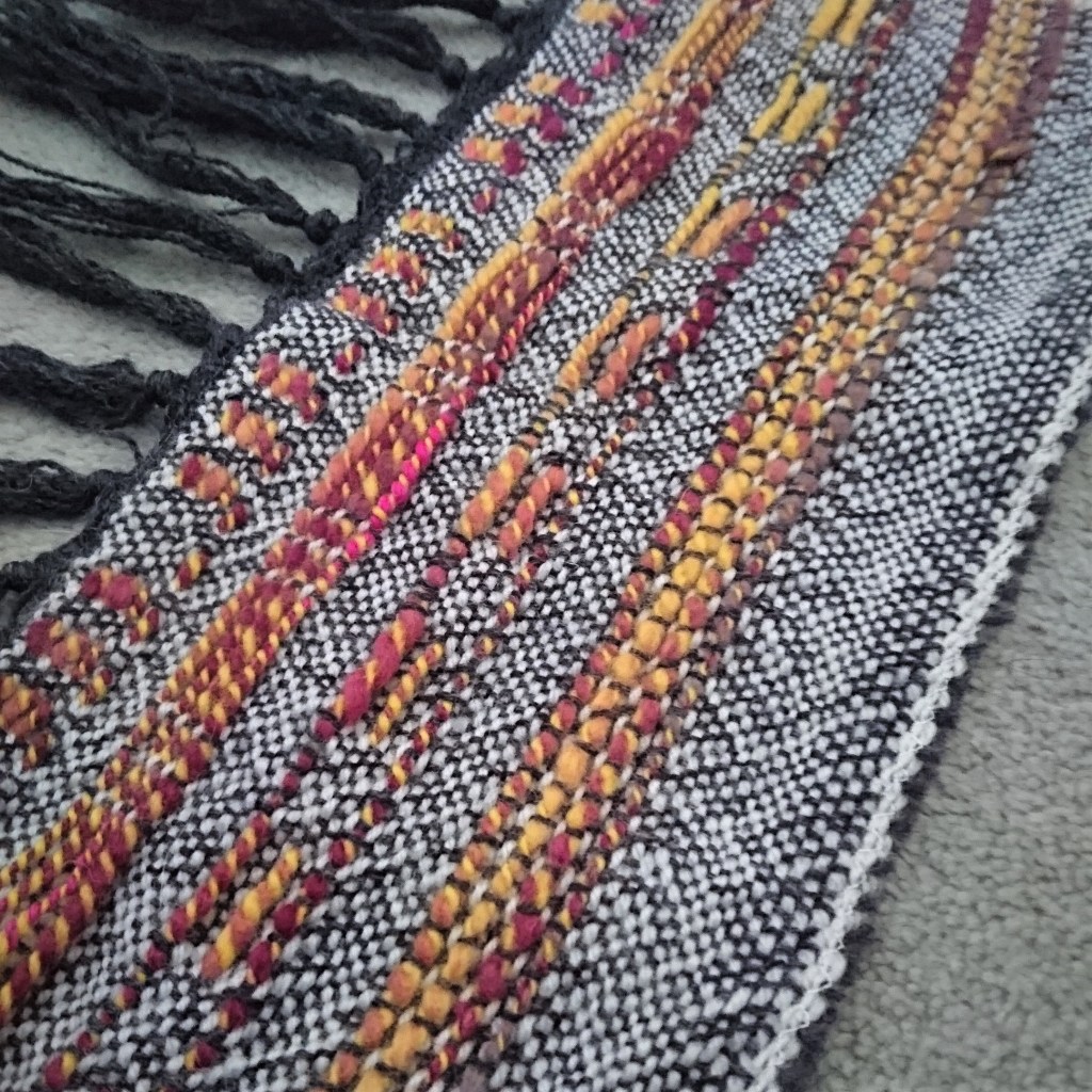 small piece of weaving laid diagonally on the carpet. Dark grey warp with white weft and random picks of bright handspun yarn in yellow, pink & purple, woven in plainweave, twill and with some large floats.