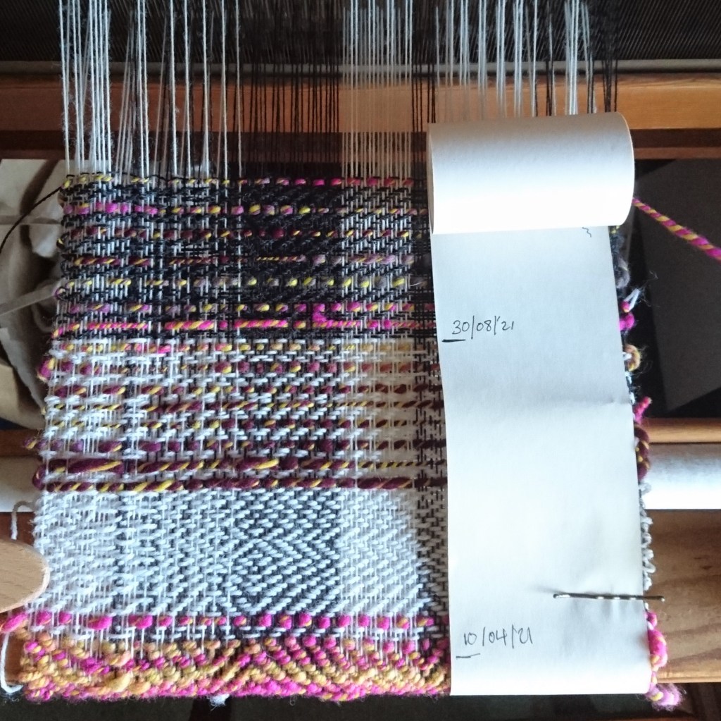 Weaving on a loom in a mix of twill patterns using grey, white thin yarns and a thick variegated handspun yarn in yellow, pink & purple. A long thing roll of paper is attached to one side with bobby pins recording the dates of weaving, it jumps from 10/04/21 to 30/08/21.